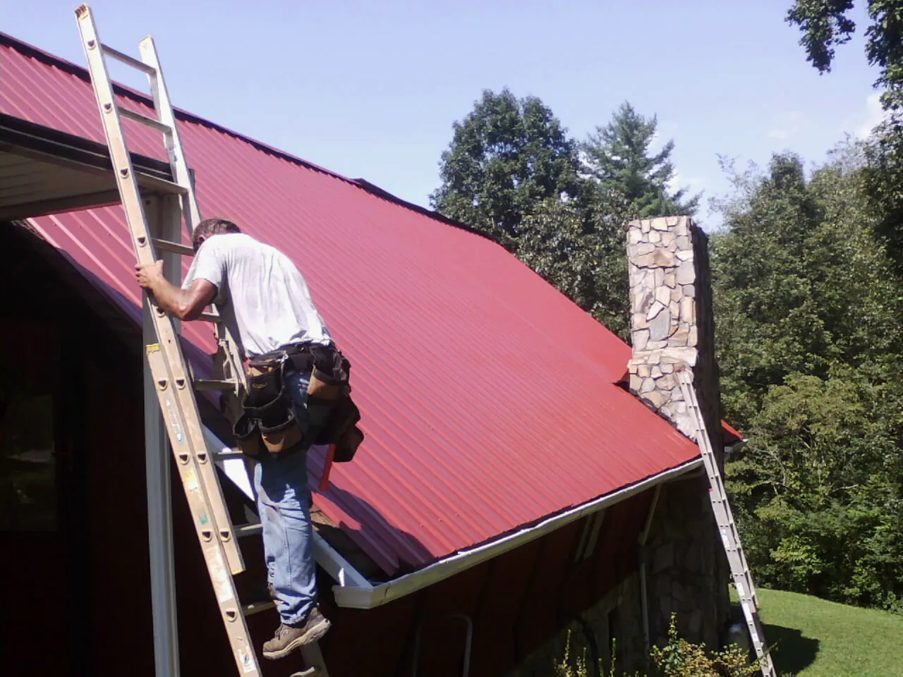 Roofer on a ladder beside a red metal roof and stone chimney — Soffit & Fascia Repair by Roofing Brothers in Collinsville